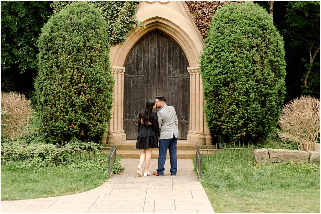 Couple kissing in front of old doors of a church with a lot of foliage around them