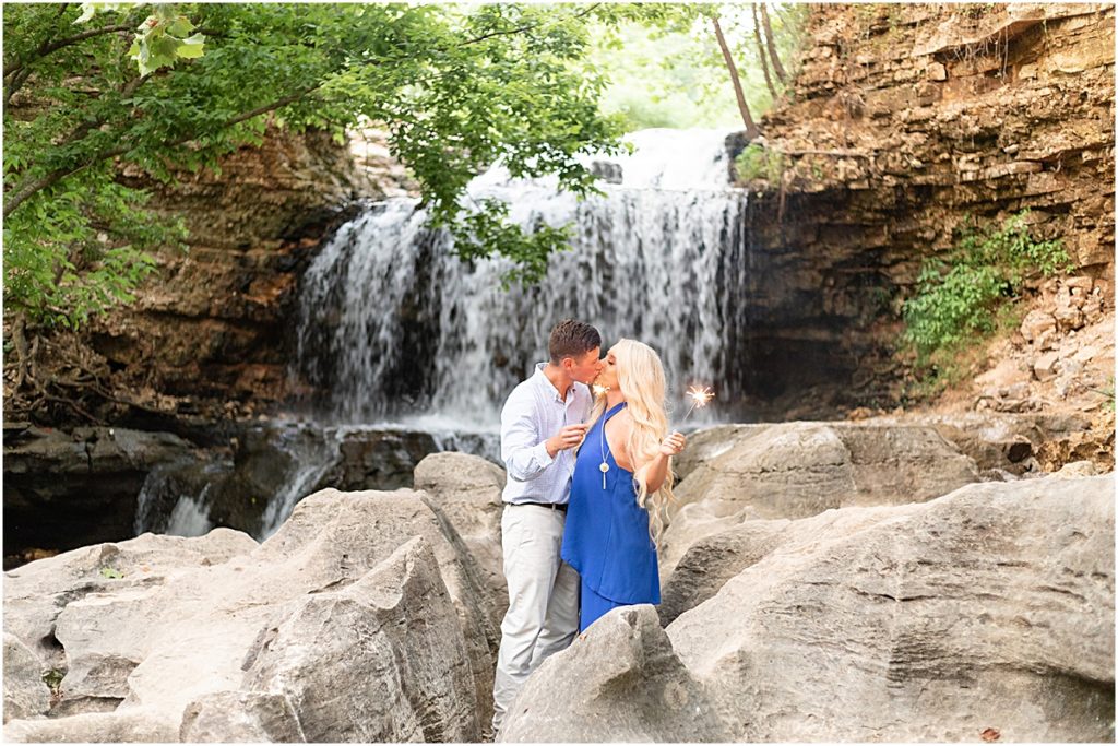 Couple standing among rocks, holding sparklers while kissing with a waterfall behind them during Engagement Photography session in NWA
