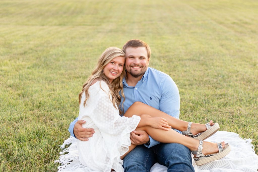 A woman sitting in her fiance's lap while he hugs her in a field during their portrait session in Northwest Arkansas.