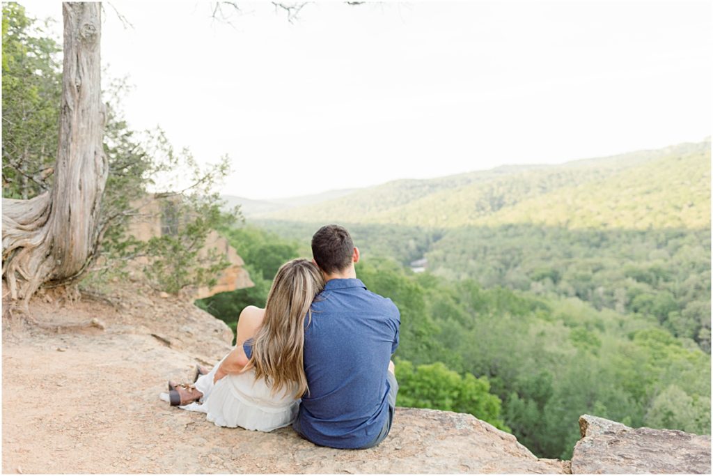 Sarah and Jarod sitting on the edge of a cliff looking out over the lush trees during their engagement session at Yellow Rock Trail.