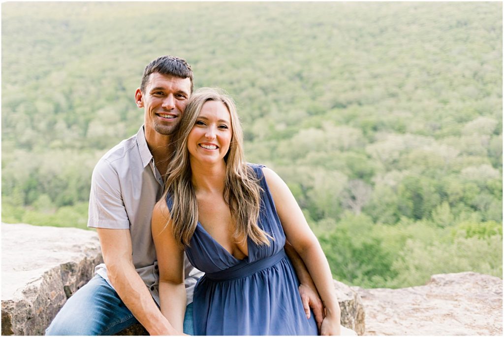 Jarod and Sarah smiling as they sit close together on the cliff's edge during their engagement session in Fayetteville, Arkansas.