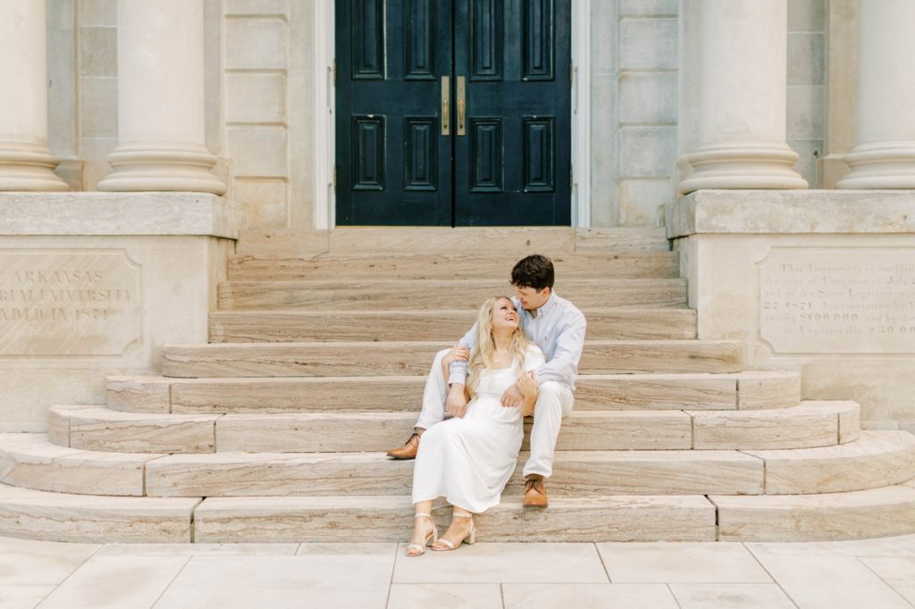 A couple sitting on the stone stairs of a building during their engagement session smiling at each other.
