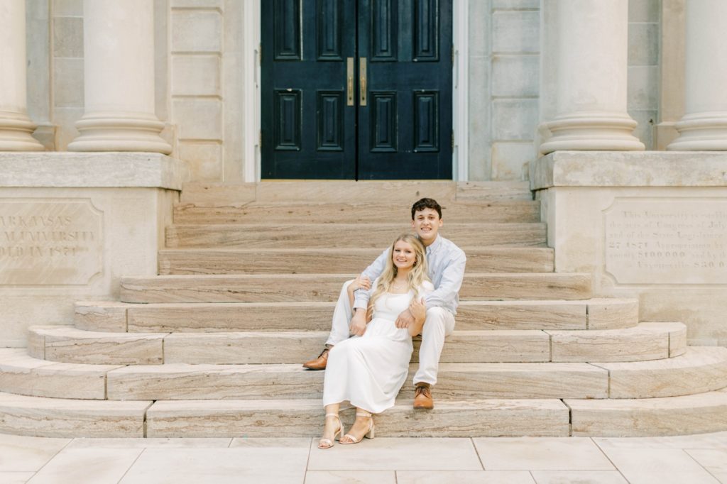 A woman sitting between her fiance's knees on the stone stairs of a building during their engagement session.