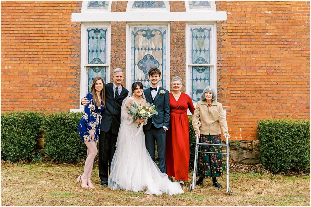 Lawrence and Emily with family outside church in front stained glass windows, brick and bushes during Arkansas Elopement 