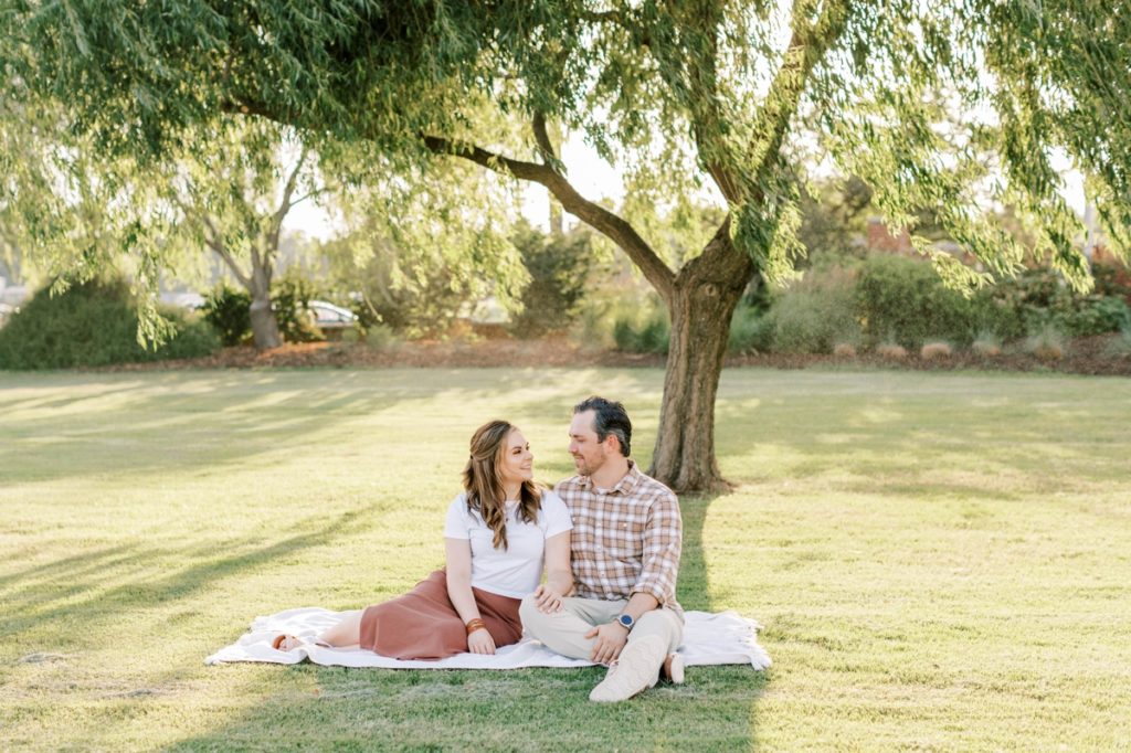 A man and woman sitting on a blanket looking lovingly at each other during their engagement session.
