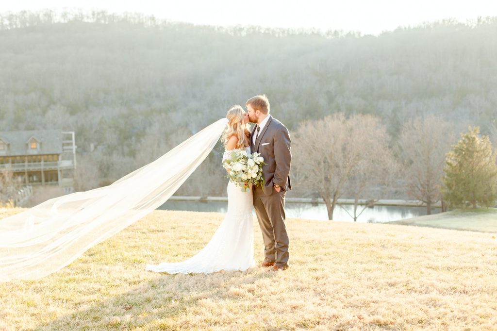 A couple kissing on a grassy hill featuring a bouquet by Carey's Flowers.