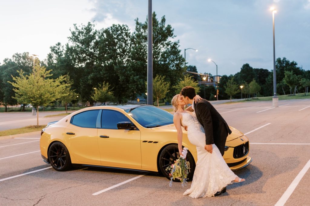 A groom dipping and kissing his bride in front of a yellow Maserati in the University of Arkansas stadium parking lot.