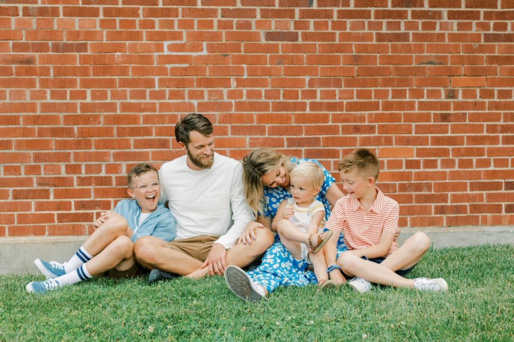 The Flake family sitting in the grass playing with each other in front of a red brick wall during their family session.