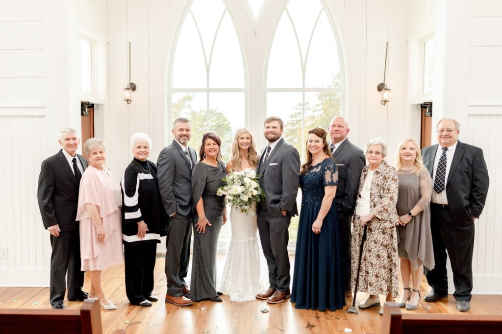 Large family photo with the bride and groom on their wedding day in front of a window filled white wall of a wedding chapel.