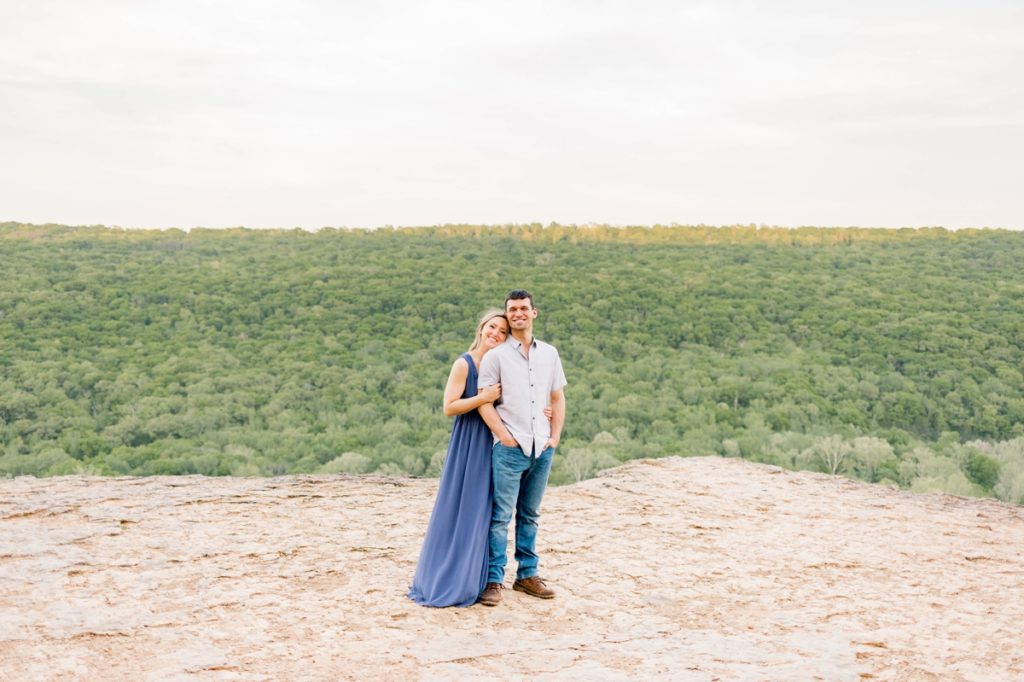A woman in a full length blue dress leaning onto her fiance's back while they smile during their engagement session.